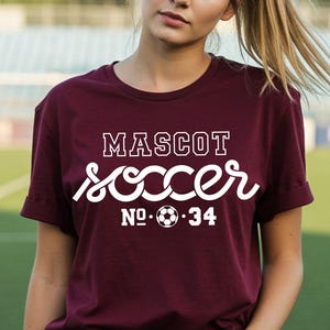 May include: A maroon t-shirt with the words "MASCOT SOCCER NO. 34" in white script and block letters. The shirt features a soccer ball graphic. The person is wearing blue jeans.