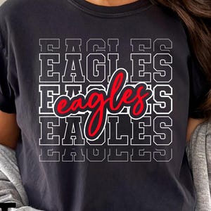 May include: Black t-shirt with the word "EAGLES" stacked in a white, block-letter design. The word "eagles" is written in red script over the stacked text. The shirt is worn with a gray cardigan.