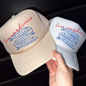 以下が含まれることがあります： ベージュと白の2つのトラッカーハット。赤い筆記体で「less sardines」の文字と、3匹の魚の青いイラストが描かれています。ハットはメッシュバックとロープのディテールが特徴です。