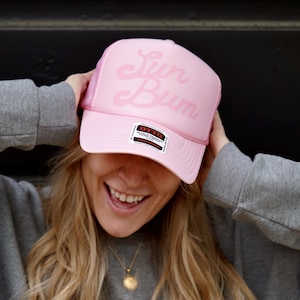 May include: A pink mesh trucker hat with the text "Sun Bum" in pink script. The hat has a black and white label with the text "Sun Bum" and "World Famous Sunscreen".
