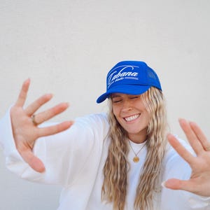 Op de afbeelding: Een vrouw draagt een blauwe trucker-pet met witte tekst die "Cabana Life" zegt. Ze glimlacht en houdt haar handen voor zich uit. Ze draagt een witte trui.