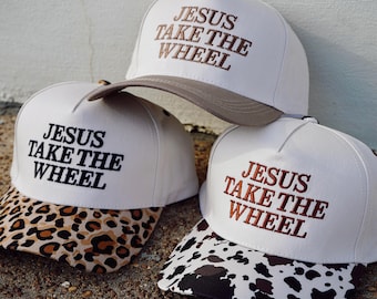 以下が含まれることがあります： 刺繍で「JESUS TAKE THE WHEEL」と書かれた3つの白い野球帽。1つは無地のベージュのつば、1つはヒョウ柄のつば、もう1つは白黒の牛柄のつばが付いています。各キャップにはしっかりとしたフロントパネルが付いています。