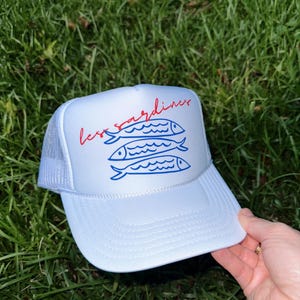 以下が含まれることがあります： カーブしたつばとメッシュのバックが付いた白いトラッカーハット。「less sardines」という赤い文字と、3匹の青い魚のイラストが描かれています。