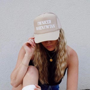 May include: A beige trucker hat with a white mesh back. The hat has the text "I'm nicer when I'm tan" printed on the front in white.