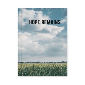 Może przedstawiać: Notatnik z czarnym napisem "HOPE REMAINS" na tle zachmurzonego nieba nad polem zielonych upraw. Okładka ma matowe wykończenie.