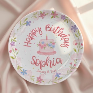 May include: A white ceramic plate with a hand-painted design. The plate features the words "Happy Birthday Sophia" in pink script, a pink cake with blue and pink flowers, and floral accents.