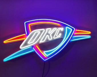 May include: Neon sign featuring the letters "OKC" in white, enclosed within a blue, red and yellow lightning bolt design. The sign is set against a purple backdrop.