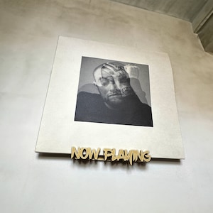 May include: A black and white portrait of a man with tattoos, holding his head, is displayed on a square, off-white background. A gold-colored metal sign that reads "NOW PLAYING" is below the portrait.