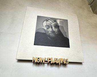 May include: A black and white portrait of a man with tattoos, holding his head, is displayed on a square, off-white background. A gold-colored metal sign that reads "NOW PLAYING" is below the portrait.