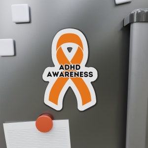 May include: Orange awareness ribbon magnet with the text "ADHD AWARENESS" on a grey refrigerator.