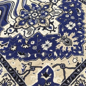 May include: Close-up of a patterned rug with a traditional design. The rug features a combination of blue, cream, and gray colors. The design includes floral and geometric patterns, creating a detailed and ornate appearance.