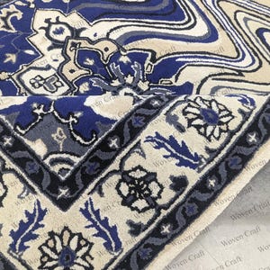 May include: A section of a blue and cream-colored rug with an intricate floral and geometric pattern. The rug's border features a dark blue design with floral motifs. The rug is partially rolled up, revealing a gray backing.