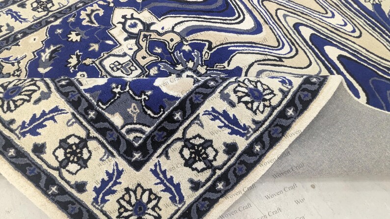 May include: A section of a blue and cream-colored rug with an intricate floral and geometric pattern. The rug's border features a dark blue design with floral motifs. The rug is partially rolled up, revealing a gray backing.