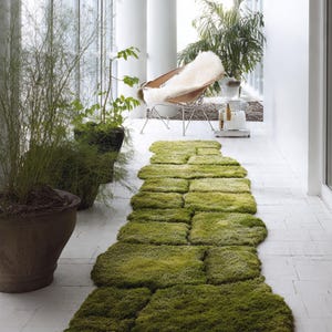 Puede incluir: Una larga alfombra verde texturizada, hecha de secciones rectangulares que se asemejan a musgo, se extiende por un pasillo embaldosado en blanco. Plantas en macetas y una silla con una piel blanca son visibles en el fondo.