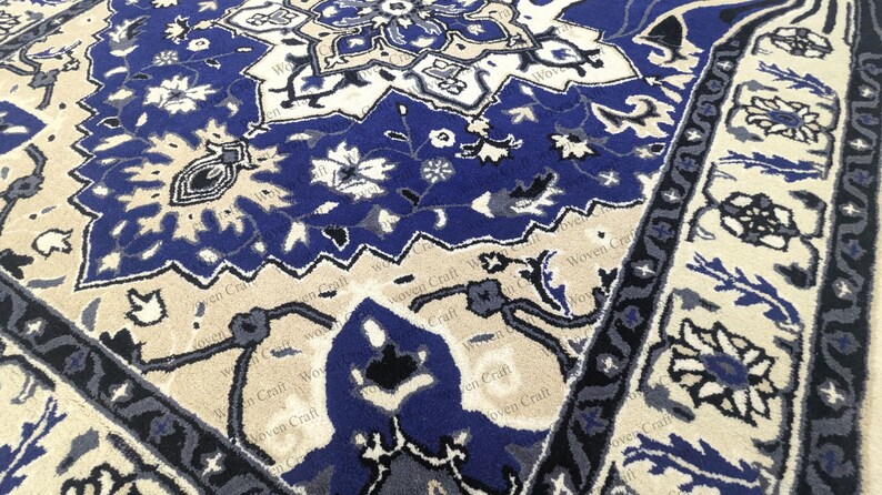 May include: Close-up of a blue and beige patterned rug. The rug features a central design with floral and geometric motifs in shades of blue, beige, and white. The border has a repeating floral pattern. The rug is made of a soft, woven material.