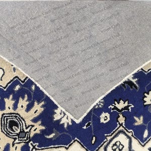 May include: A section of a blue, beige, and gray patterned rug with a floral design. The rug's underside is visible, showing a gray backing with the text "Woven Craft" repeated across it. The rug has a traditional style.