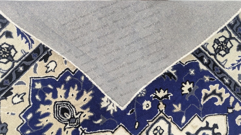 May include: A section of a blue, beige, and gray patterned rug with a floral design. The rug's underside is visible, showing a gray backing with the text "Woven Craft" repeated across it. The rug has a traditional style.