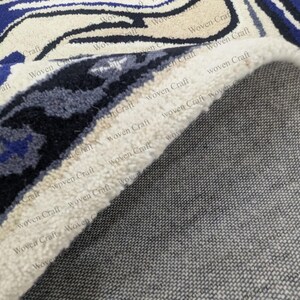 May include: Close-up of a rolled-up area rug with a detailed pattern. The rug features a cream-colored background with blue and black floral designs. The underside of the rug is a gray woven fabric.