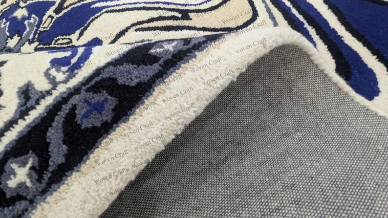 May include: Close-up of a rolled-up area rug with a detailed pattern. The rug features a cream-colored background with blue and black floral designs. The underside of the rug is a gray woven fabric.