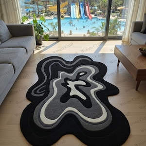 Könnte beinhalten: Abstrakter Teppich mit fließendem Design in Schwarz, Grau und Weiß. Der Teppich hat ein geschichtetes Muster. Er liegt auf einem hellen Holzboden und verleiht dem Raum einen Hauch von modernem Stil. Die Abmessungen des Teppichs sind in Zentimetern.