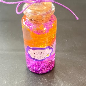 May include: A small glass bottle with a cork stopper and a purple ribbon tied around the top. The bottle is filled with orange glitter and a purple label that says "Travel Potion".
