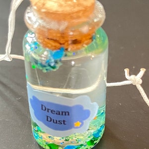 May include: A small glass bottle with a cork stopper filled with green and blue glitter. The bottle has a blue label with the text "Dream Dust" and a small yellow star.