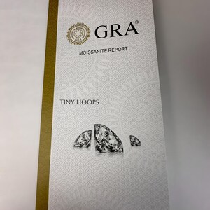 Op de afbeelding: Een wit en gouden moissanite-rapport met de tekst "GRA Moissanite Report" en "Tiny Hoops". Het rapport bevat een logo met de tekst "GRA" en een afbeelding van drie diamanten.