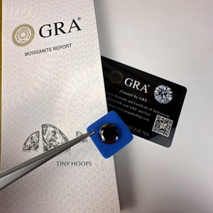 Op de afbeelding: Een close-up van een zwarte moissanite-edelsteen in een blauw schuimvierkant. De edelsteen wordt vastgehouden door een pincet. De afbeelding toont ook een GRA Moissanite Report-kaart met de tekst "GRA Moissanite Report" en "Gemaakt door GRA".