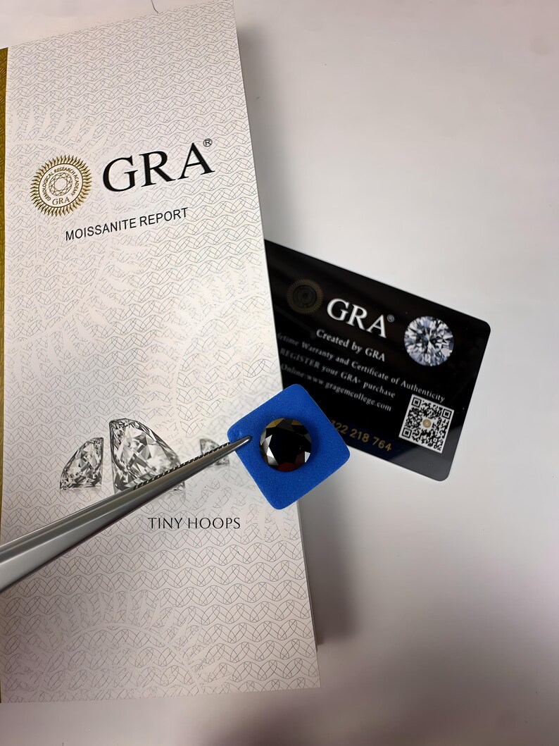 Op de afbeelding: Een close-up van een zwarte moissanite-edelsteen in een blauw schuimvierkant. De edelsteen wordt vastgehouden door een pincet. De afbeelding toont ook een GRA Moissanite Report-kaart met de tekst "GRA Moissanite Report" en "Gemaakt door GRA".