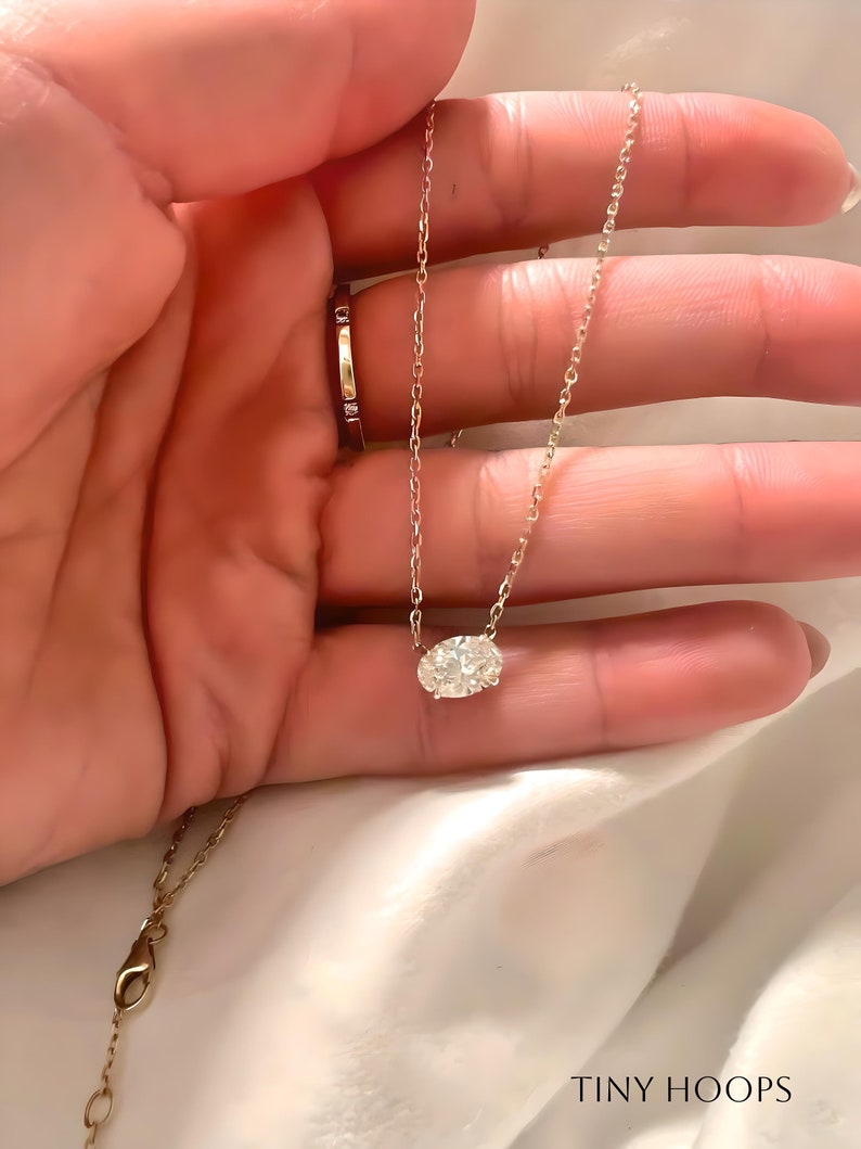 May include: A delicate gold chain necklace with a single oval-shaped diamond pendant. The necklace is displayed on a hand, with the diamond pendant centered in the palm.