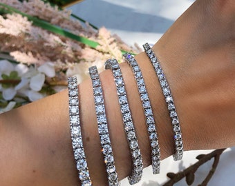 Könnte beinhalten: Fünf silberne Tennisarmbänder mit klaren Zirkonia-Steinen. Die Armbänder sind auf dem Handgelenk einer Person übereinander gelegt.