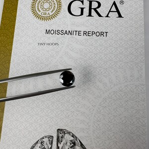 Op de afbeelding: Een witte kaart met de tekst "GRA Moissanite Report" en "Tiny Hoops". Een zwarte edelsteen wordt in een pincet boven de kaart gehouden. De kaart heeft een patroon van overlappende cirkels.