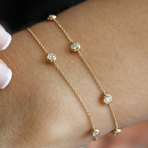 May include: Two delicate gold bracelets with small round diamonds. The bracelets are layered on a person's wrist.