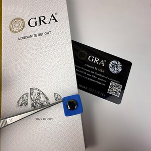Op de afbeelding: Een witte kaart met de tekst "GRA Moissanite Report" en een klein diamantvormig beeld. De kaart ligt naast een blauw vierkant met een zwarte diamantvormige steen erin.