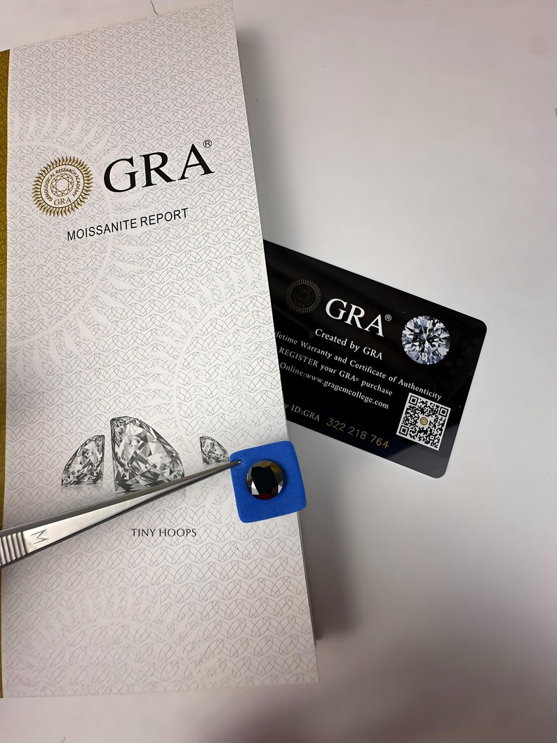 Op de afbeelding: Een witte kaart met de tekst "GRA Moissanite Report" en een klein diamantvormig beeld. De kaart ligt naast een blauw vierkant met een zwarte diamantvormige steen erin.