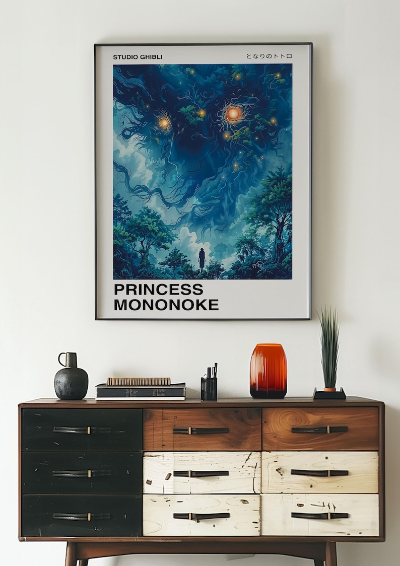 May include: A framed poster of the Studio Ghibli film Princess Mononoke. The poster features a dark blue and green illustration of a forest spirit with glowing eyes. The text "Studio Ghibli" and "Princess Mononoke" is printed in black at the top and bottom of the poster. The poster is hanging on a white wall above a wooden dresser.