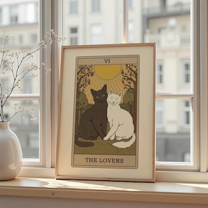 May include: A framed tarot card print featuring two cats, one black and one white, sitting together. The card is labeled "VI THE LOVERS" and depicts a sun, trees, and fruit. The print has a beige border and is displayed on a windowsill.