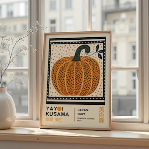 Peut inclure: Impression d'art encadrée représentant une citrouille orange stylisée avec des points noirs et une tige verte. L'œuvre est placée sur un fond crème avec des points orange et une bordure noire. Le texte comprend "YAYOI KUSAMA" et "JAPAN 1997".