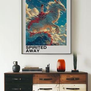 May include: A framed poster with a colorful illustration of a white dragon with a red tail, carrying a young girl on its back. The poster has the text "Studio Ghibli" and "Spirited Away" in black text. The Japanese text "となりのトトロ" is also on the poster.
