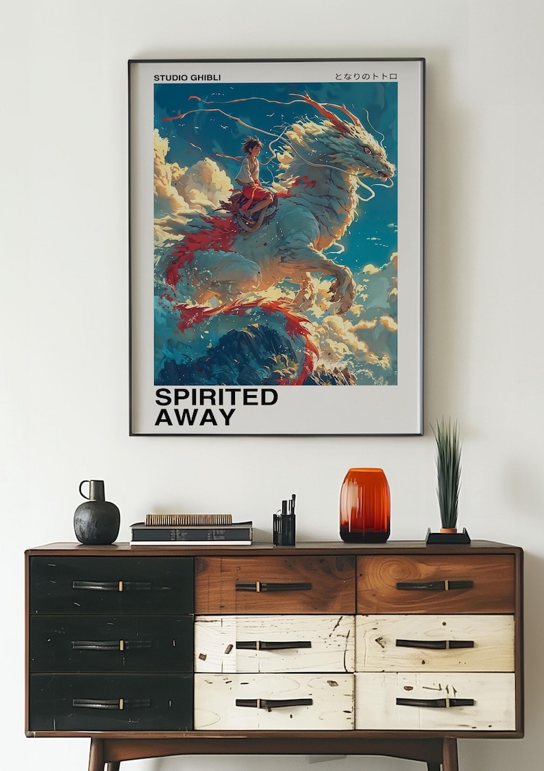 May include: A framed poster with a colorful illustration of a white dragon with a red tail, carrying a young girl on its back. The poster has the text "Studio Ghibli" and "Spirited Away" in black text. The Japanese text "となりのトトロ" is also on the poster.