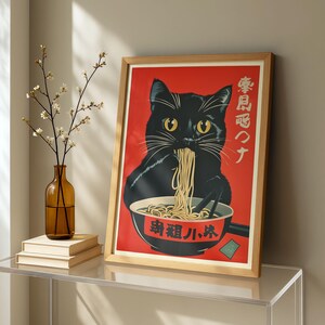 May include: A framed art print featuring a black cat eating noodles from a bowl, set against a red background. The cat has yellow eyes and is the central focus. The print has Japanese text and is in a wooden frame.