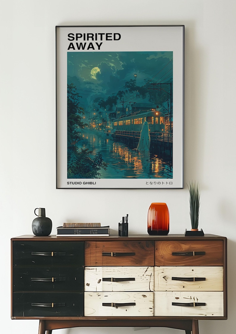 May include: A framed poster of the Studio Ghibli film "Spirited Away" with the Japanese title "となりのトトロ" and the text "Studio Ghibli" at the bottom. The poster features a nighttime scene with a train, a river, and a glowing figure.