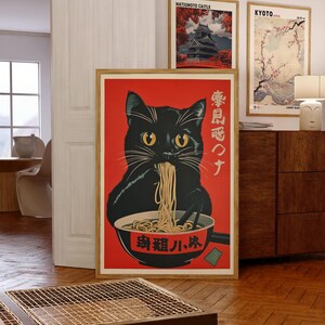 May include: A framed art print featuring a black cat eating ramen from a bowl. The cat has yellow eyes and is set against a red background. Japanese text is visible on the print. Two other framed prints are visible in the background.