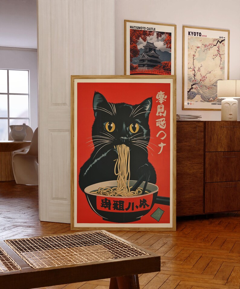 May include: A framed art print featuring a black cat eating ramen from a bowl. The cat has yellow eyes and is set against a red background. Japanese text is visible on the print. Two other framed prints are visible in the background.