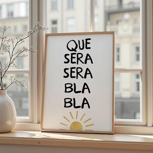 Op de afbeelding: Een ingelijste print met de woorden "QUE SERA SERA BLA BLA" in het zwart, met een gouden zonillustratie aan de onderkant. De print zit in een licht houten frame en staat op een vensterbank.