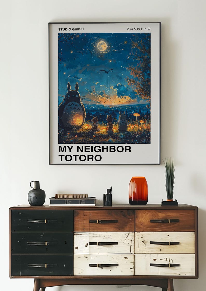 May include: A framed poster of the Studio Ghibli film "My Neighbor Totoro" featuring the titular character Totoro and two young girls sitting in front of a glowing moon and a cityscape in the distance. The poster is hanging on a white wall above a wooden dresser with drawers.