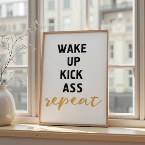 Op de afbeelding: Een ingelijste print met de woorden "WAKE UP KICK ASS REPEAT" in zwarte en gouden letters. De print wordt weergegeven op een vensterbank naast een witte vaas met een bloemstuk.