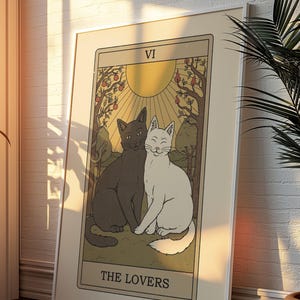 May include: A framed tarot card print featuring two cats, one black and one white, sitting together. The card is labeled "THE LOVERS" and depicts a sun, trees, and fruit. The card is in a beige frame.