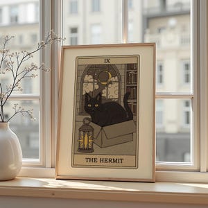May include: A framed tarot card print featuring a black cat in a box, with a nighttime library scene in the background. The card reads "THE HERMIT" at the bottom, with the Roman numeral "IX" at the top. A lantern is in the foreground.