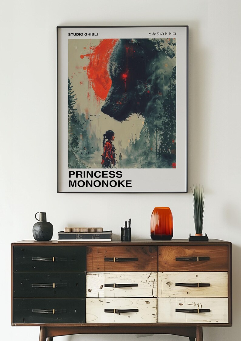 May include: A framed poster with a black and red illustration of a wolf and a young woman. The poster has the text "Studio Ghibli" and "Princess Mononoke" in black text. The poster is hanging on a white wall above a wooden dresser.